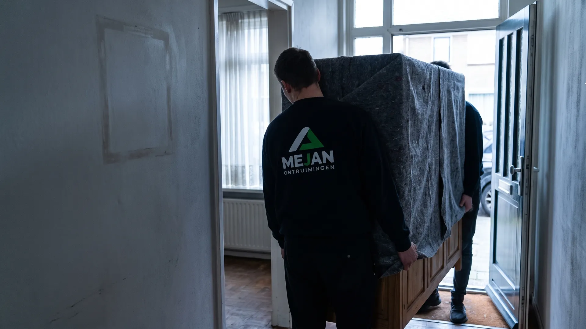 Twee medewerkers van Mejan dragen een in dekens gepakte kast door de gang naar buiten