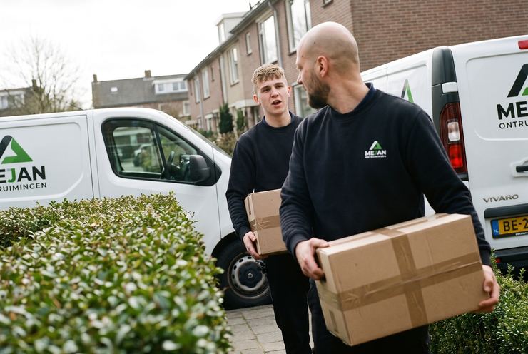 Twee medewerkers lopen met dozen over het tuinpad naar de bus