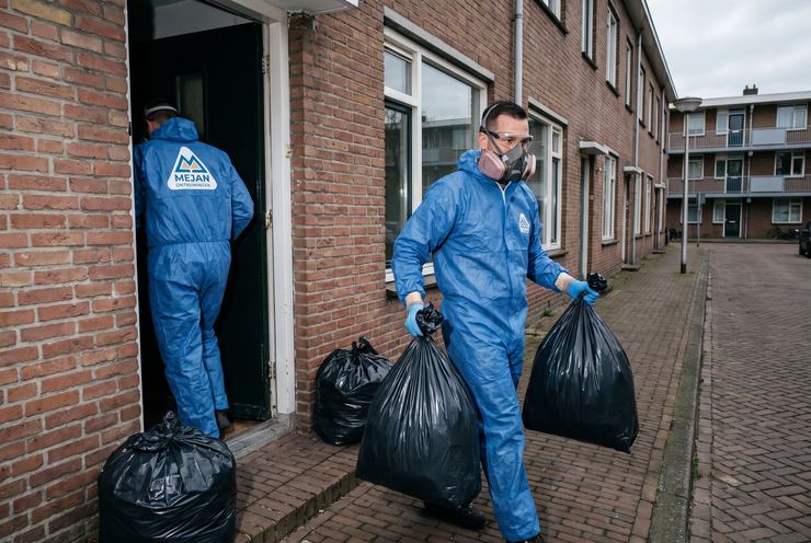 Medewerkers in beschermende kleding dragen afvalzakken uit een woning naar buiten